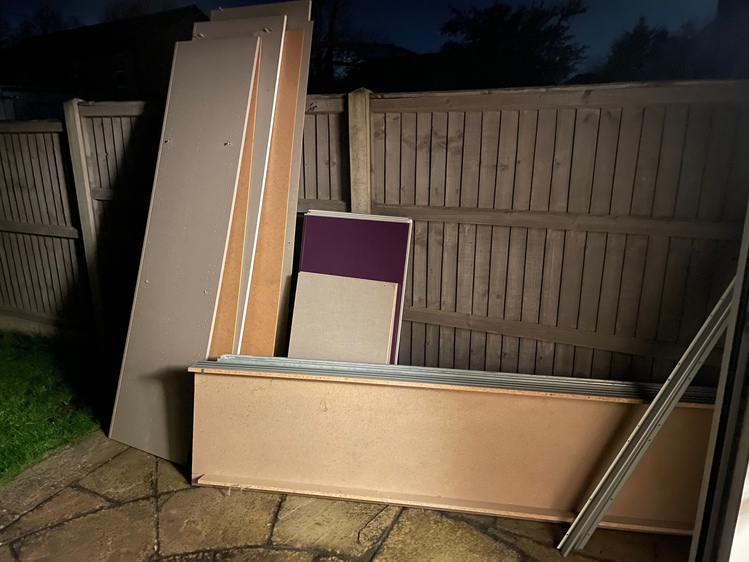 A pile of flat-pack wardrobe pieces stacked against a fence.